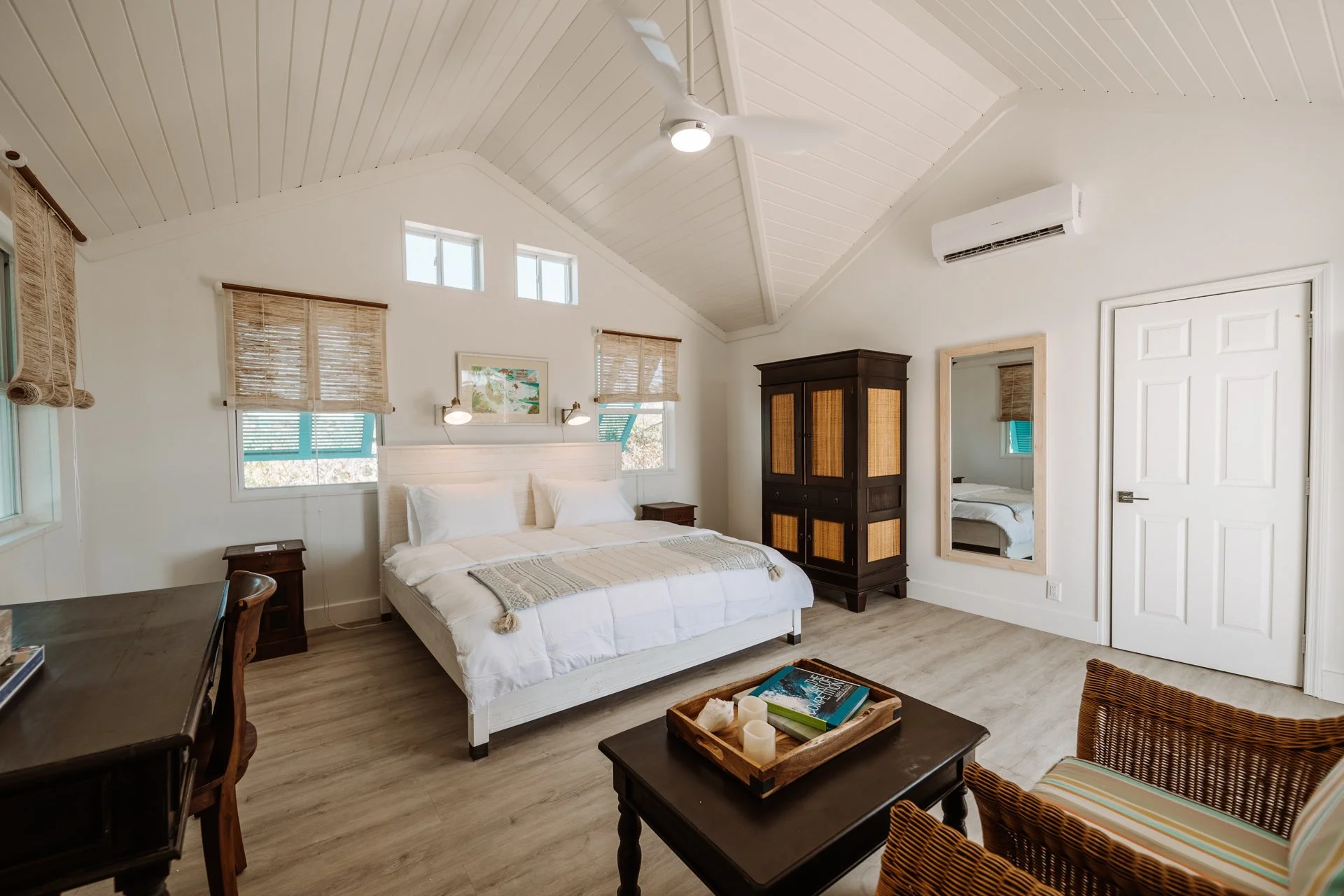 A bedroom with a white bed, wooden furniture, a mirror, windows with blinds, and a ceiling fan.