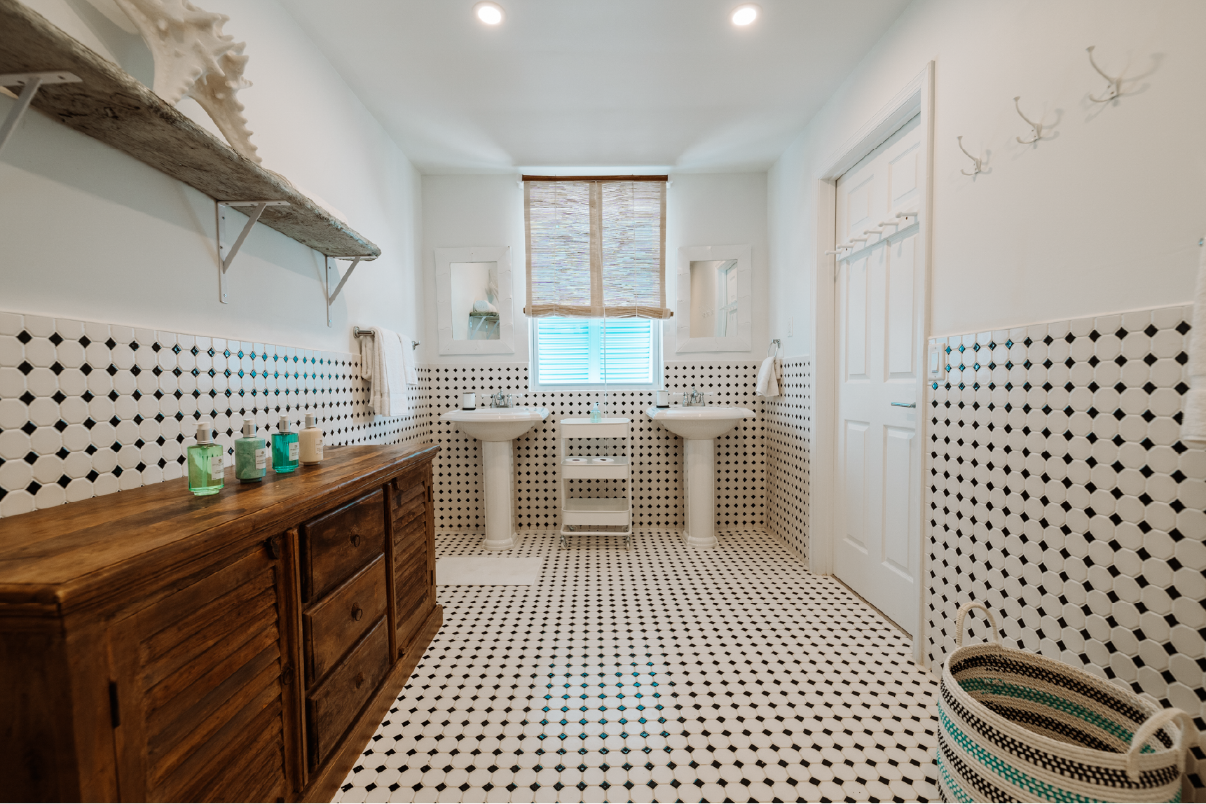 A spacious bathroom with black and white hexagonal tile throughout, two pedestal sinks below a window with blinds, a wooden cabinet on the left, and a striped basket on the right.