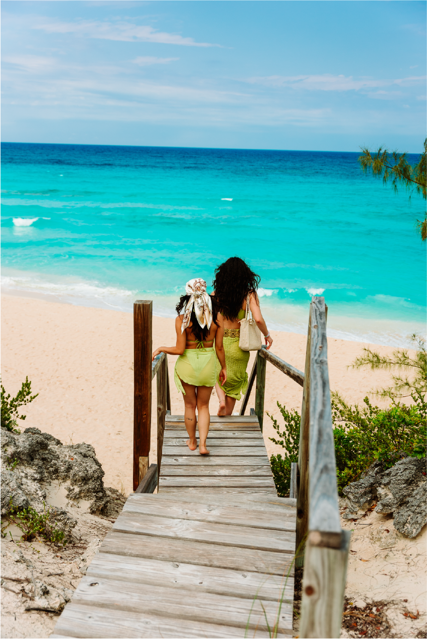 Two women in summer dresses walking down a wooden staircase onto a sandy beach with turquoise ocean water and a partly cloudy sky.