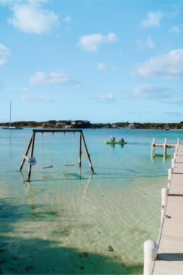 Calm ocean with a wooden swing set partially submerged, a small dock, and a kayak with two people paddling, under a partly cloudy blue sky.
