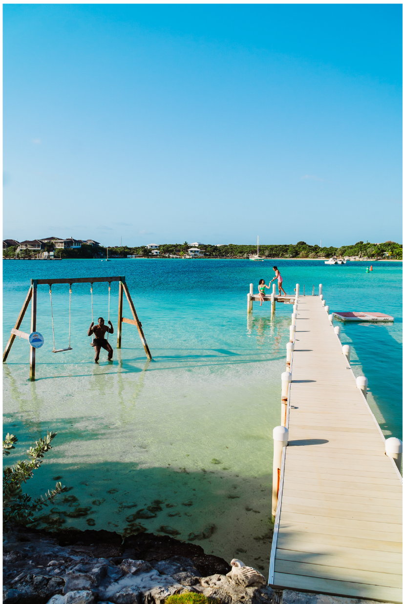 People enjoying a day at a beach with a floating dock, a swing set in shallow water, and clear blue sky and water.