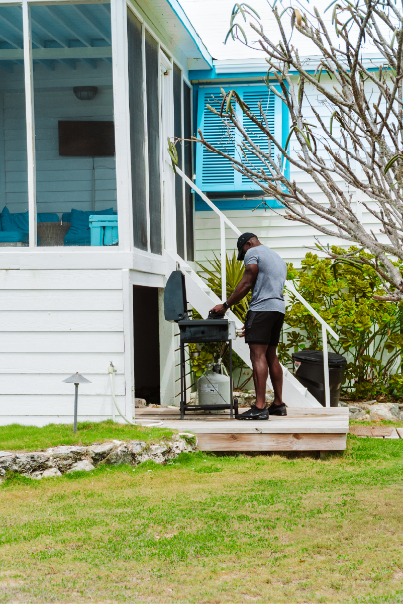 A man wearing a gray t-shirt, black shorts, and a black cap grilling outdoors on a small barbecue grill next to a house with white siding and blue window shutters, surrounded by greenery.