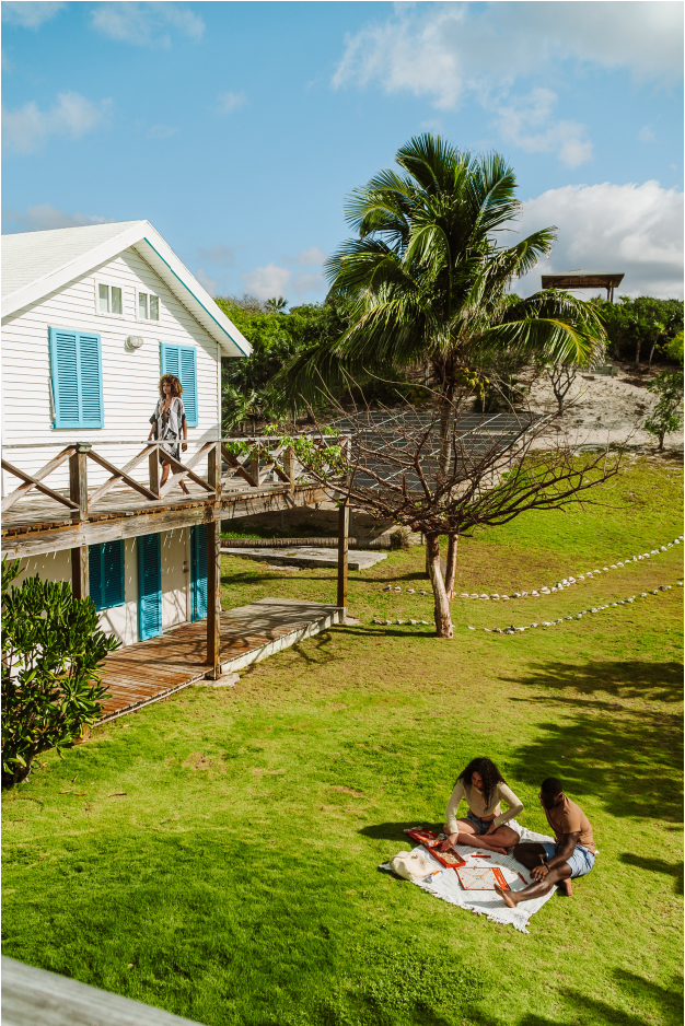 Two people sitting on a blanket on the grass, playing a game and enjoying a sunny day outside a white house with blue shutters and a wooden deck. A woman is walking on the deck and a large palm tree is nearby.