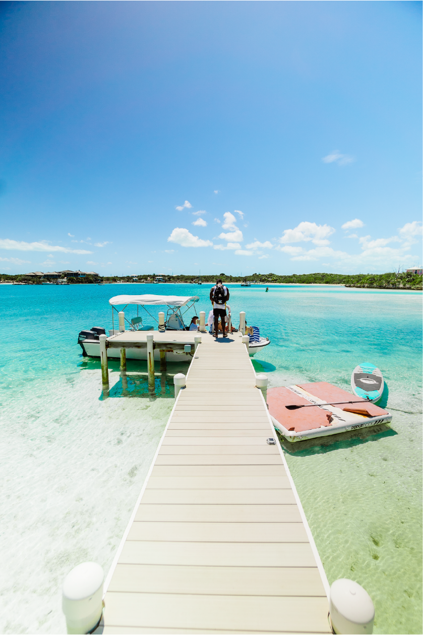A person standing on a dock leading out to a boat at a tropical beach with clear turquoise water, a paddleboard, and a cloudy blue sky.