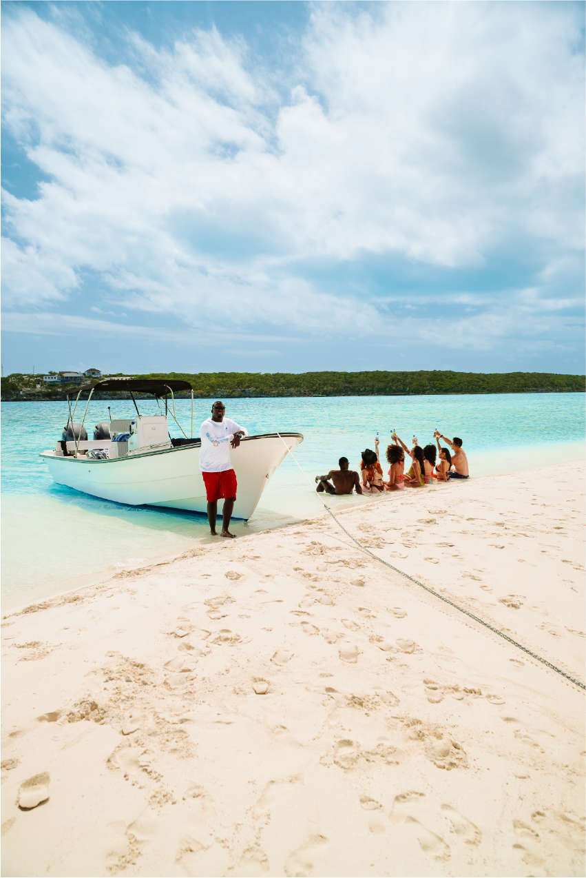 A group of people sitting on a beach near a boat on clear water, with a man standing in front of the boat and holding a rope, under a partly cloudy sky.