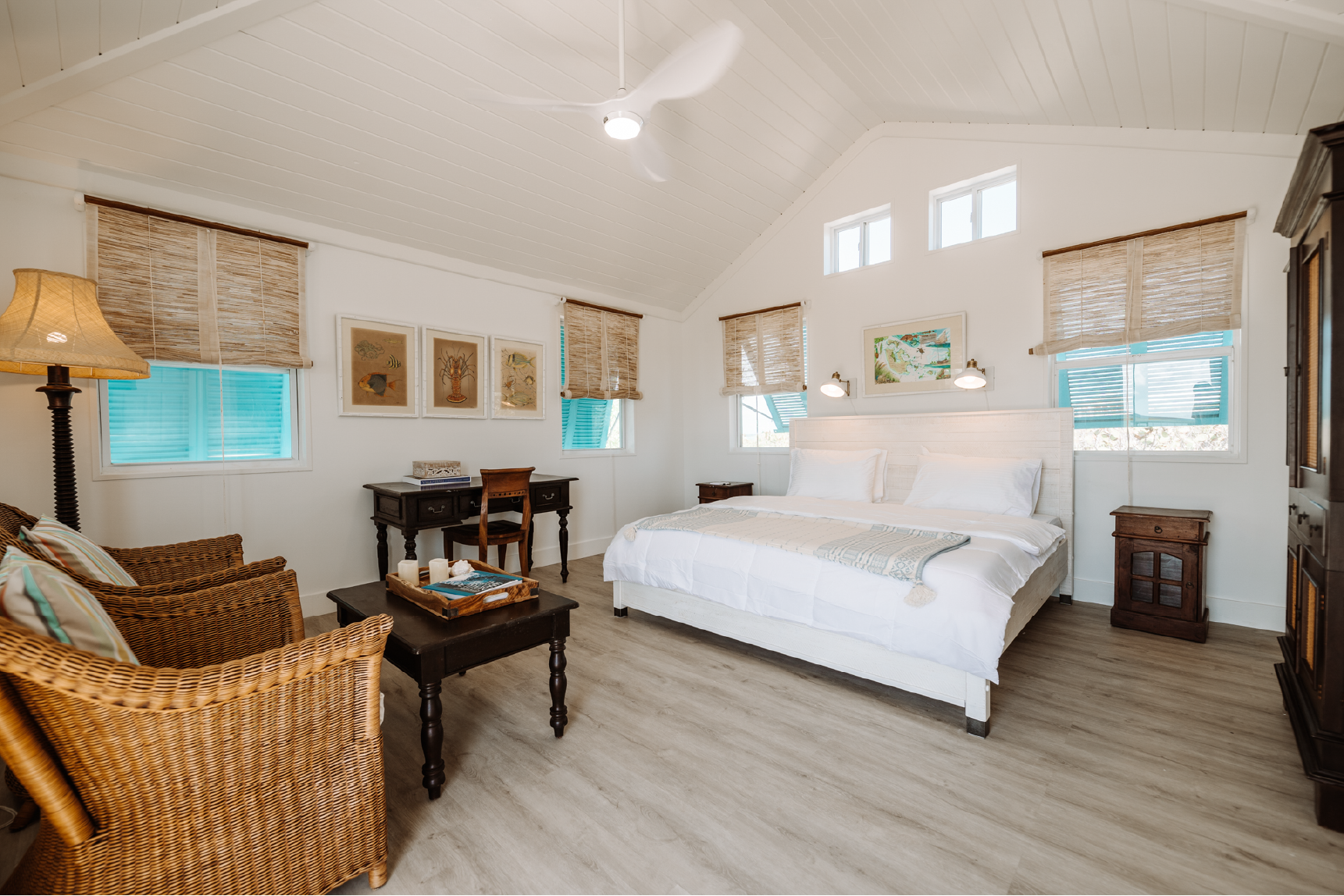 Bright, airy bedroom with white walls, sloped ceiling, and multiple windows with woven blinds. Features a white bed with a headboard, small side tables, a desk, wicker chairs, and coastal artwork.