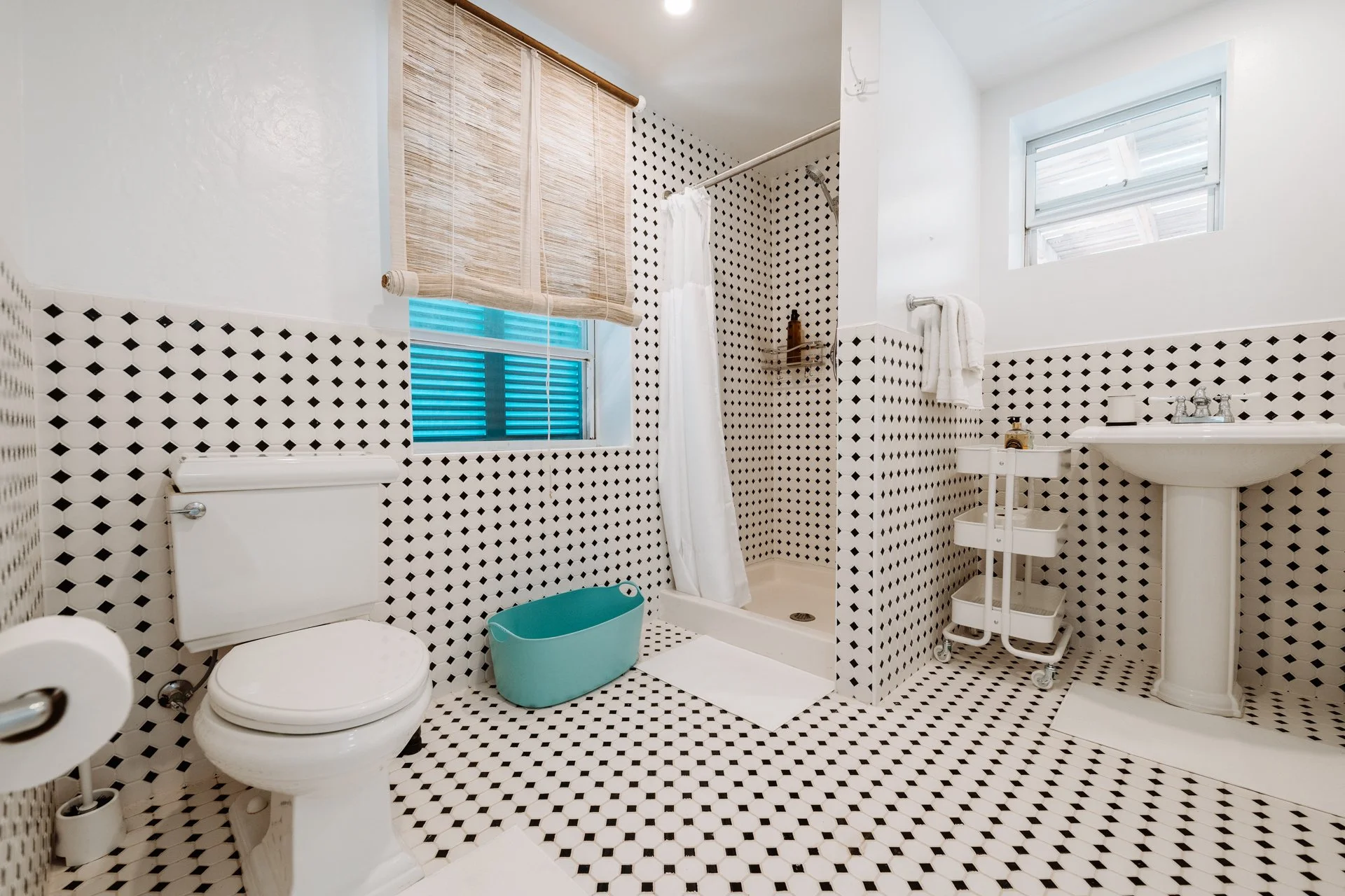 A bathroom with white and black patterned tiles on the walls and floor. It features a toilet, a small bathtub with a shower curtain, a pedestal sink, a window with a bamboo shade, and a small rolling cart with toiletries.
