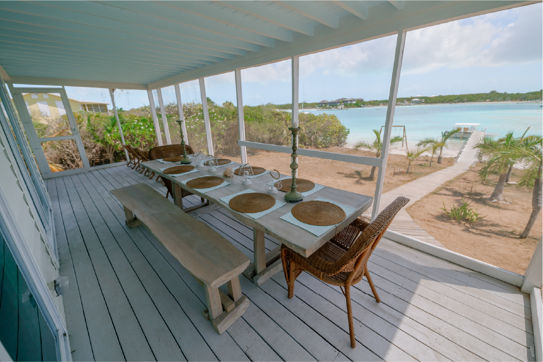 A dining table set with placemats, plates, and candles on a porch overlooking a beach, with a sandy pathway, palm trees, and calm ocean in the background.