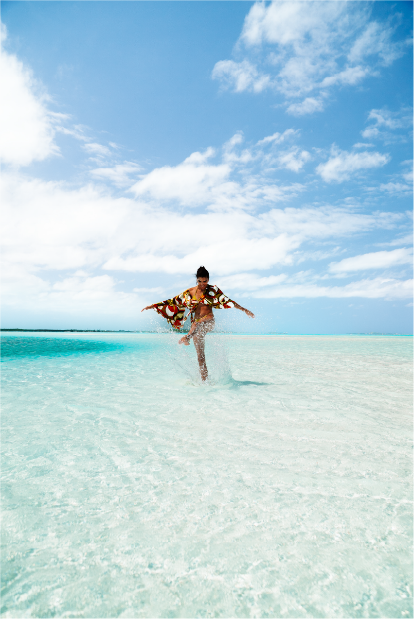 A woman in a colorful cover-up enjoying a splash in shallow clear turquoise water on a sunny beach under a blue sky with white clouds.
