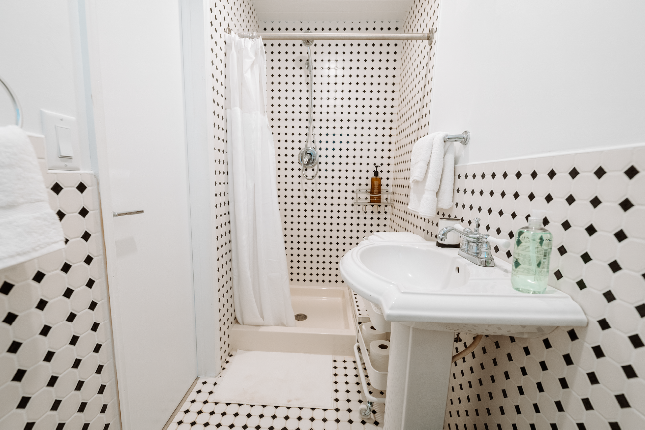 Small bathroom with black-and-white hexagon tile pattern on walls and floor, shower area with white curtain, pedestal sink, towel rack with towel, soap dispenser, and toilet paper holder.