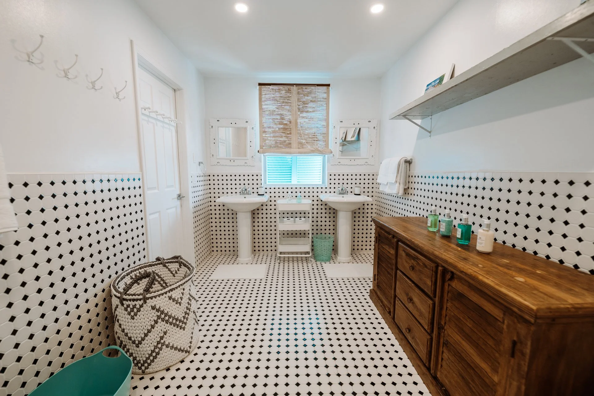 Bathroom with white walls, black and white patterned tile floor, and two sinks with mirrors. There is a wooden cabinet with bottles on top, a woven basket, and a small green trash can.