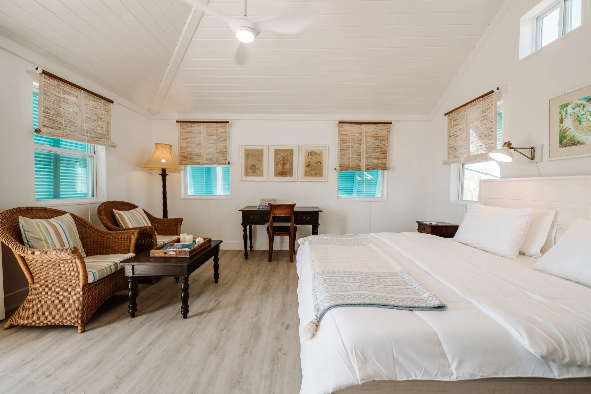 A bright, airy bedroom with a large white bed, side tables, wicker armchairs, a small desk, and various windows with woven pink blinds.