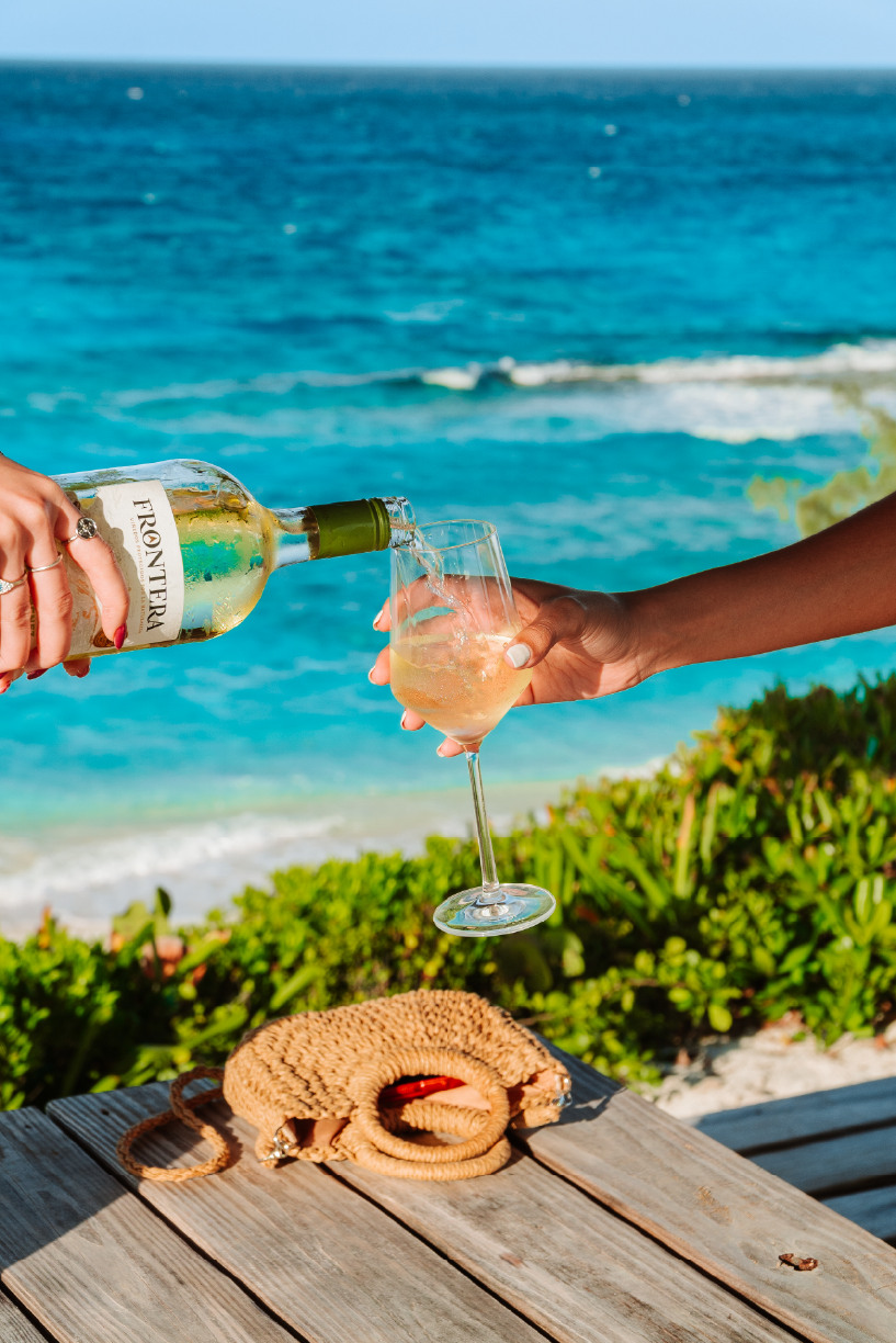 People at the beach pouring wine into a glass with a ocean view in the background, on a wooden table with a beige purse.