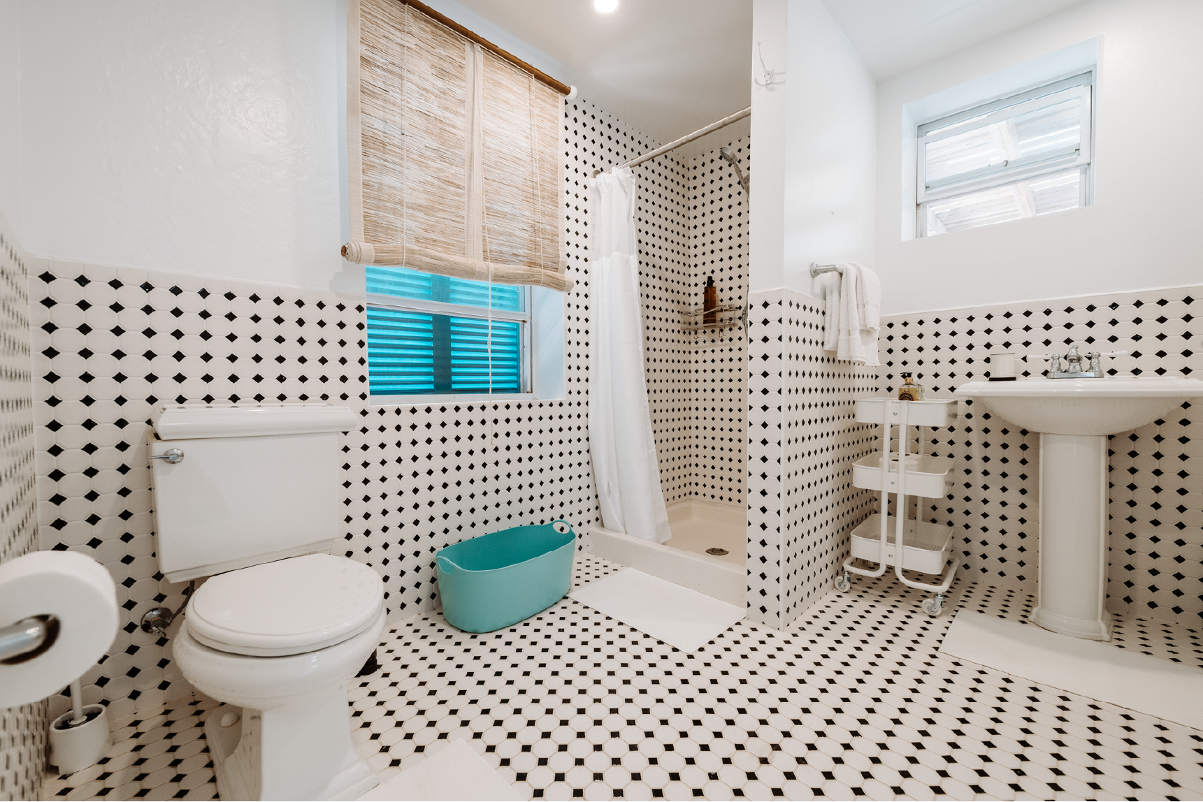 Bathroom with black and white patterned tiles, containing a toilet, a shower area with a white curtain, a small teal tub, a window with a bamboo shade, a white pedestal sink, a rolled towel on a rack, and a small rolling cart.