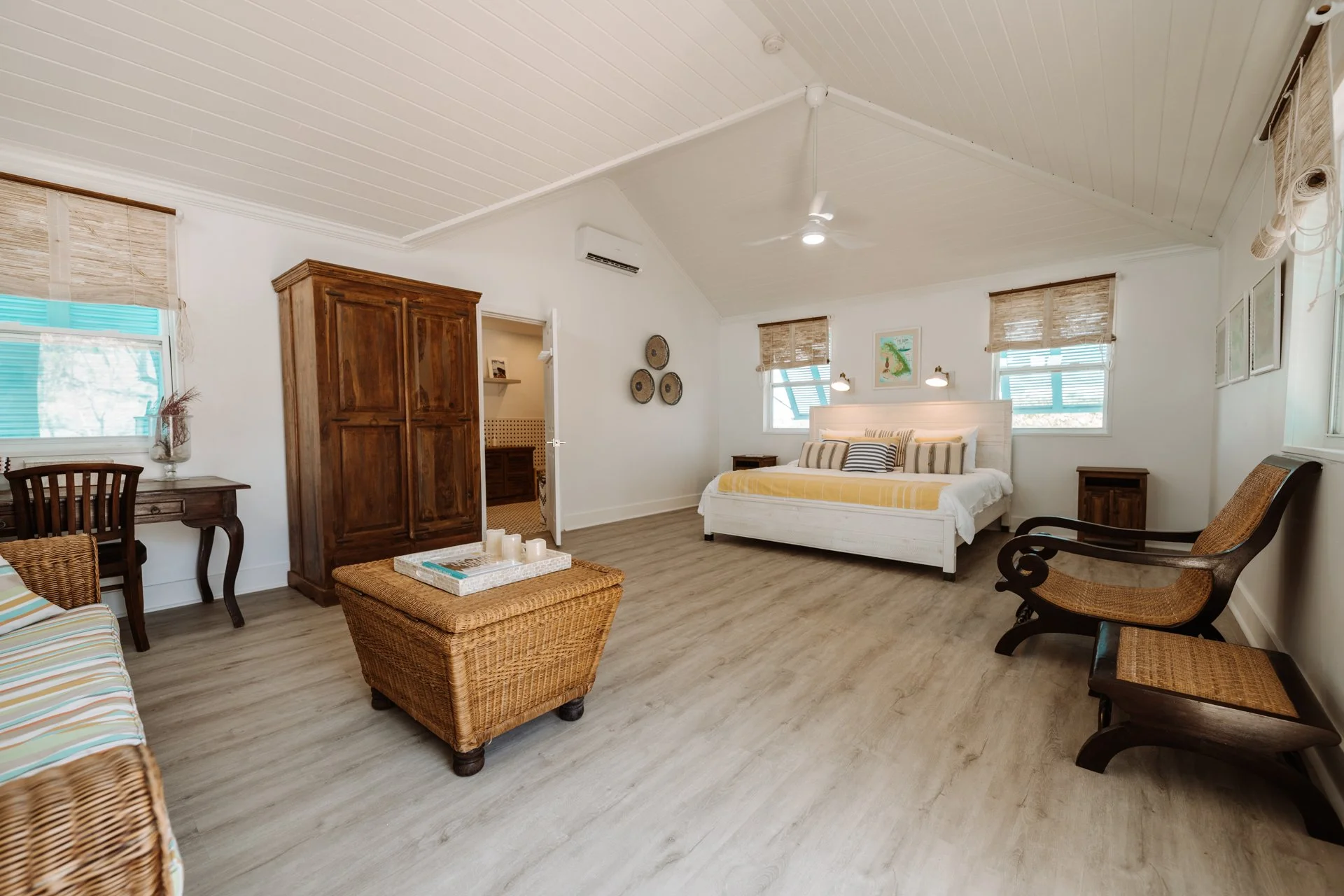 Bright bedroom with white walls, light wood flooring, and a vaulted ceiling. It has a white bed with striped pillows, two small wooden nightstands, a woven armchair, a wicker ottoman with candles, a striped sofa, and a wooden wardrobe. There are thre