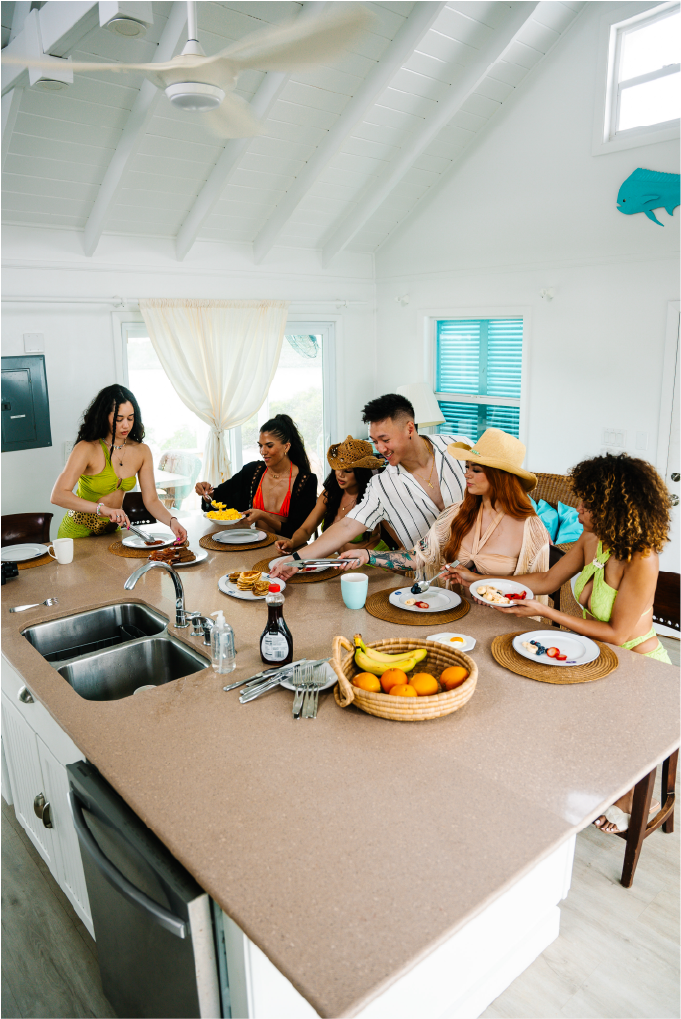 Group of friends enjoying breakfast in a bright, airy kitchen, with some serving food, and others eating, surrounded by kitchen utensils, fruit, and breakfast dishes.