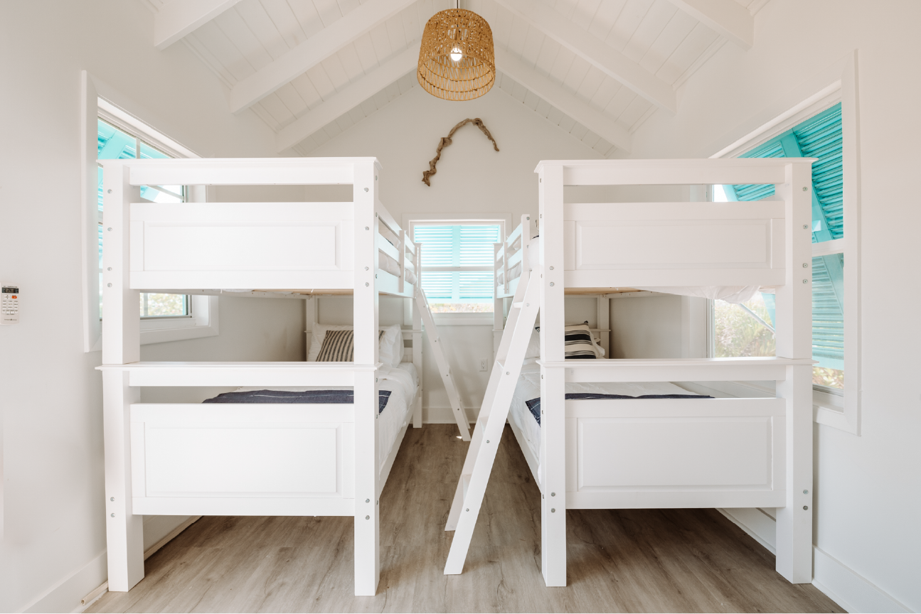 Room with two white bunk beds, each with a ladder, and striped pillows, windows with blue shutters, wood flooring, wicker lampshade hanging from ceiling, and a piece of driftwood decor on the wall.