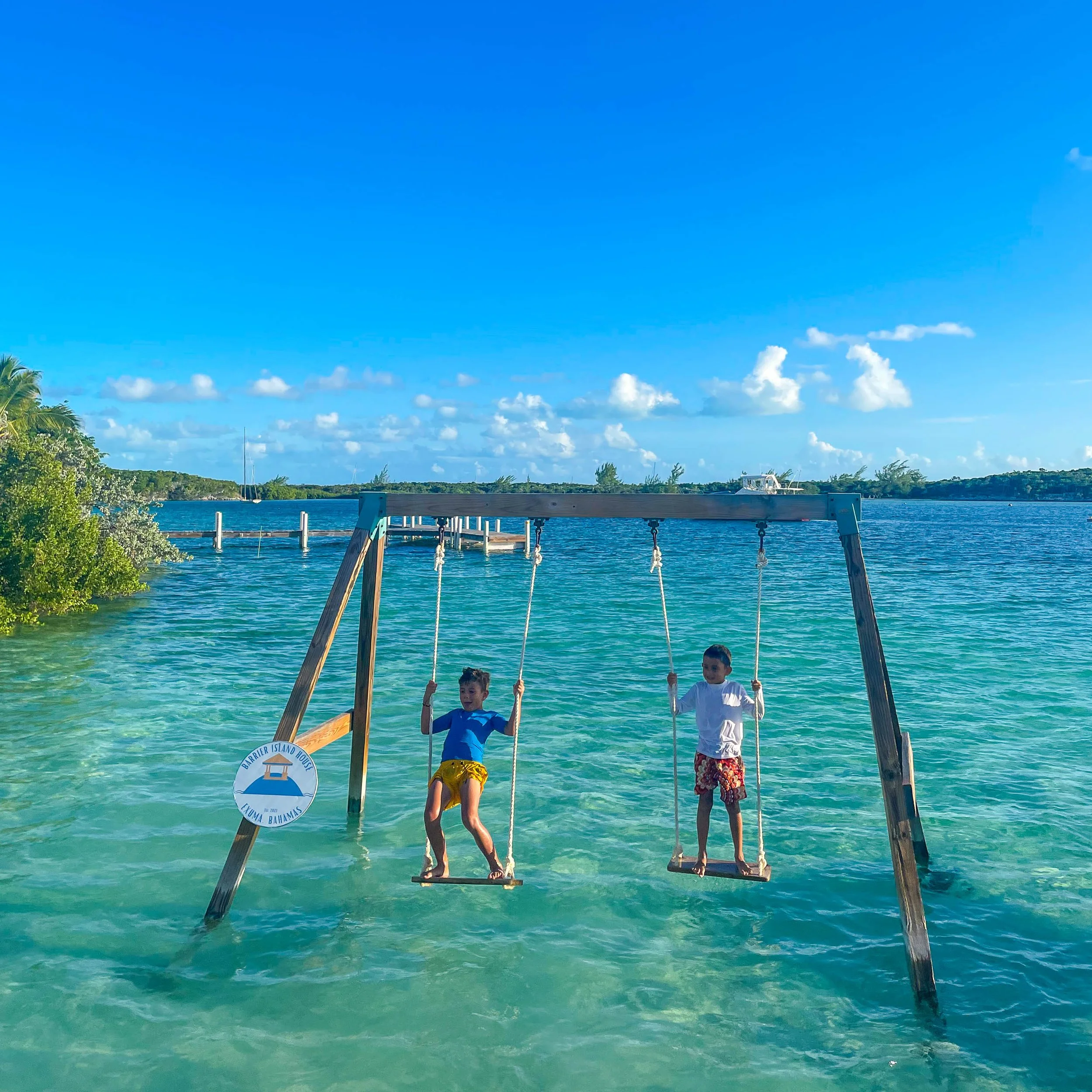Two children playing on swings over shallow, clear turquoise water with a dock and boat in the background under a sunny blue sky with scattered clouds.