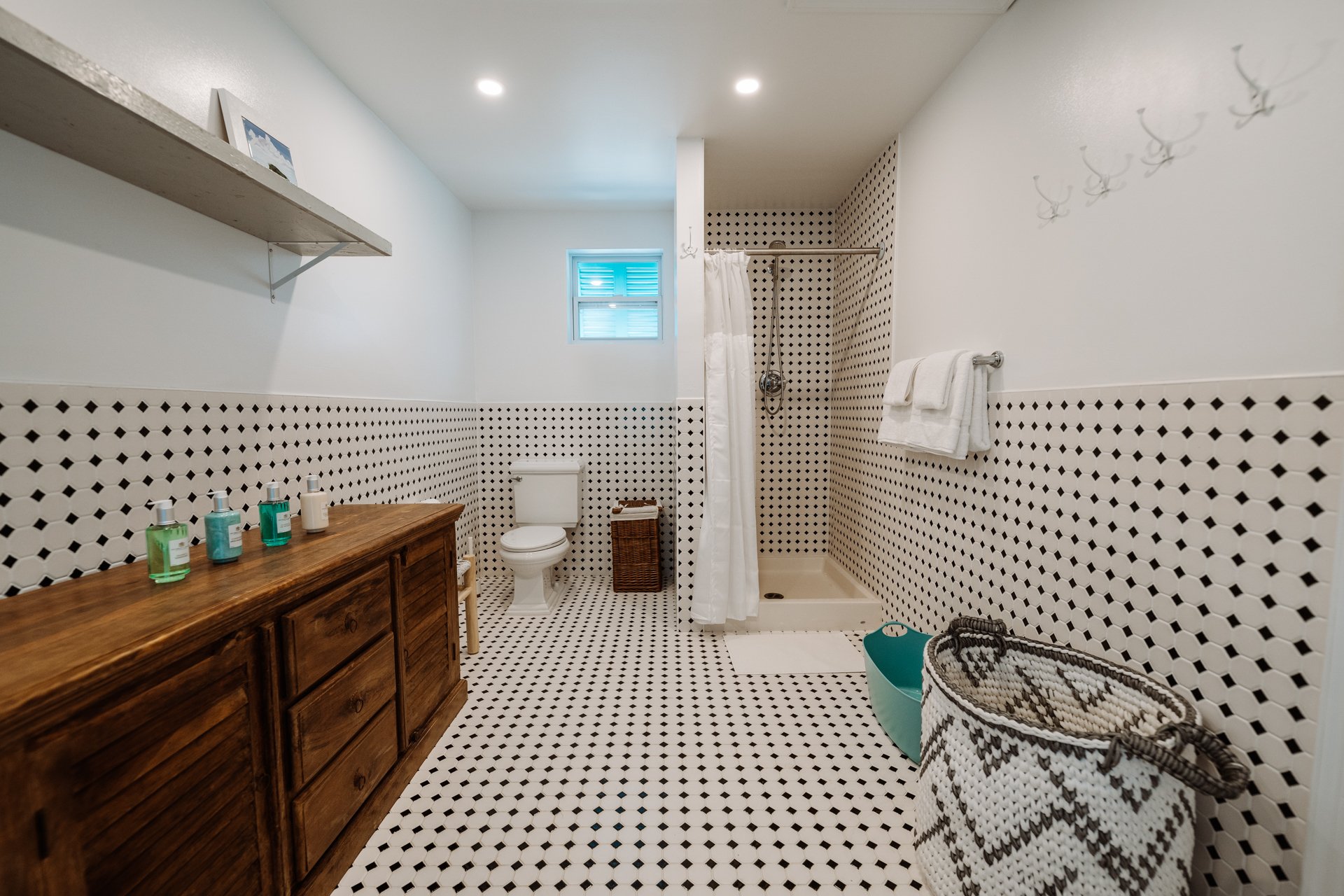 Bathroom with black and white patterned tiles, a wooden cabinet with toiletries, a toilet, a small window, a shower with a curtain, and storage baskets.