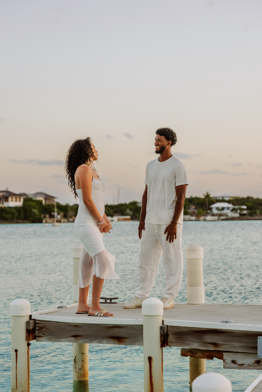 A couple standing on a dock by the water, smiling and facing each other during sunset.