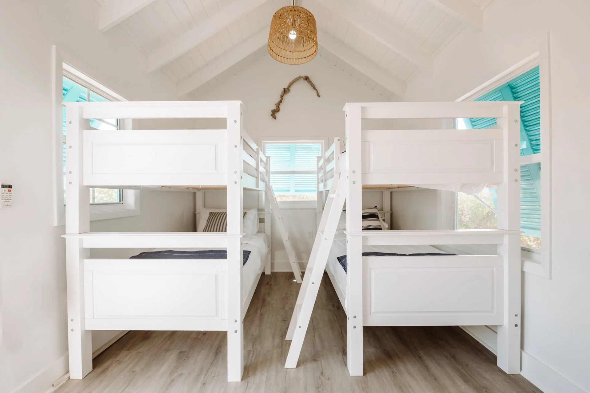 A bedroom with four white bunk beds arranged in a square, with two on each side. The beds are stacked with a ladder leading up to the top bunks. The room has white walls, a vaulted ceiling with exposed beams, a woven rattan light fixture, a piece of 