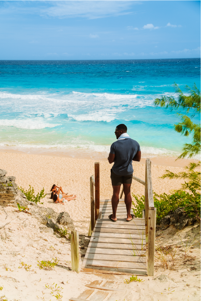 Man standing on a wooden staircase looking at a beach with two women sitting on the sand, ocean waves, and sky in the background.