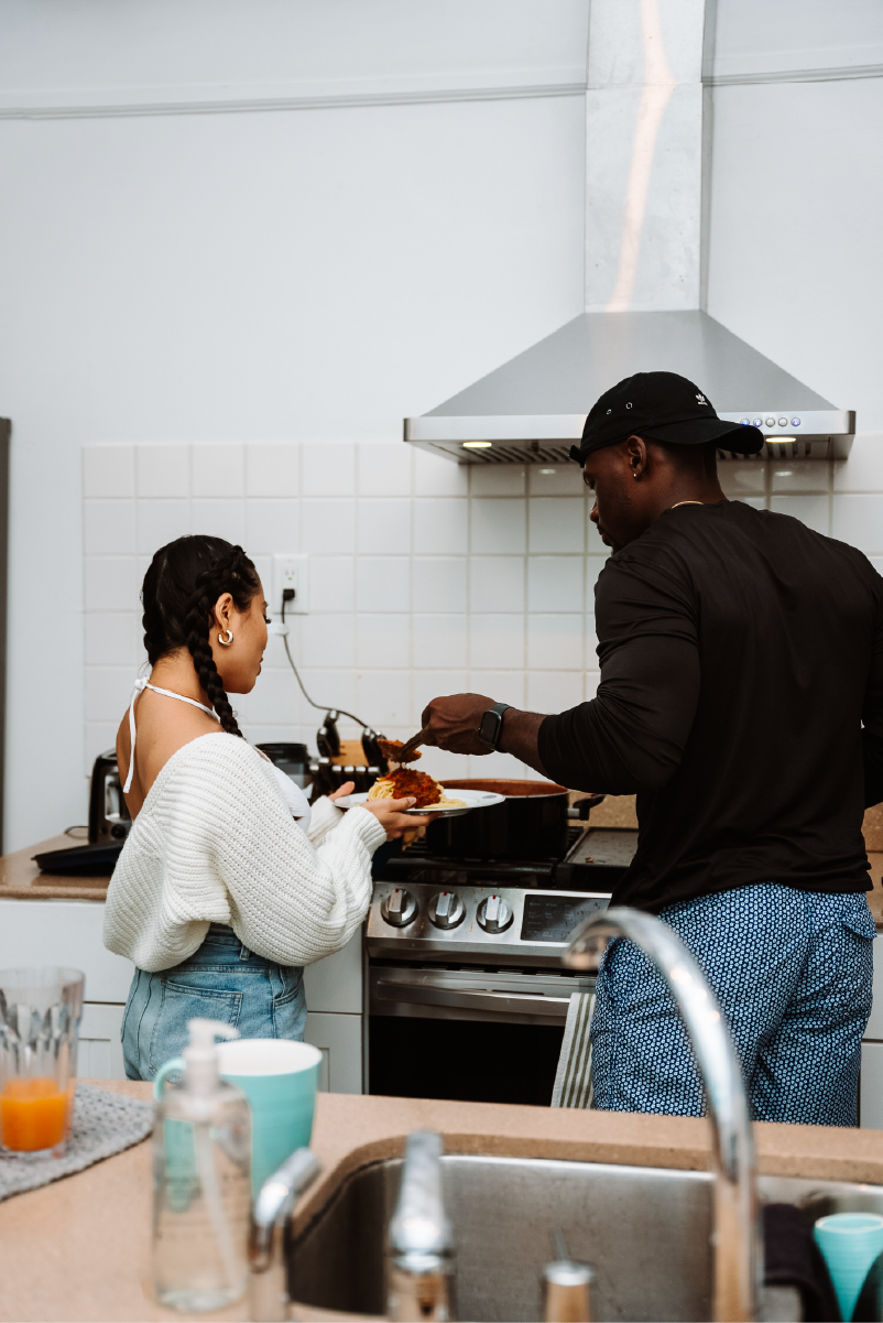 A man and a woman cooking in a modern kitchen. The man is serving food onto a plate held by the woman. The kitchen has a stove, white tile backsplash, and a range hood.