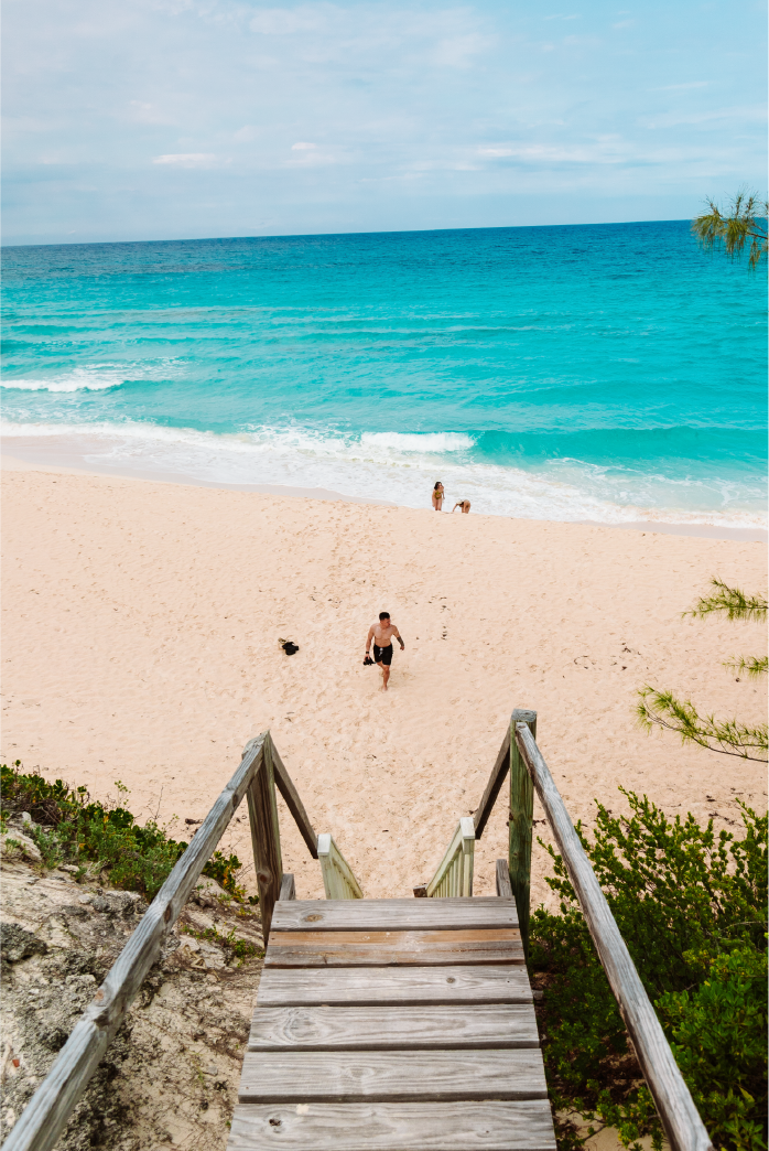 Wooden stairs leading down to a sandy beach with a man walking, two people by the shoreline, and clear blue ocean in the background