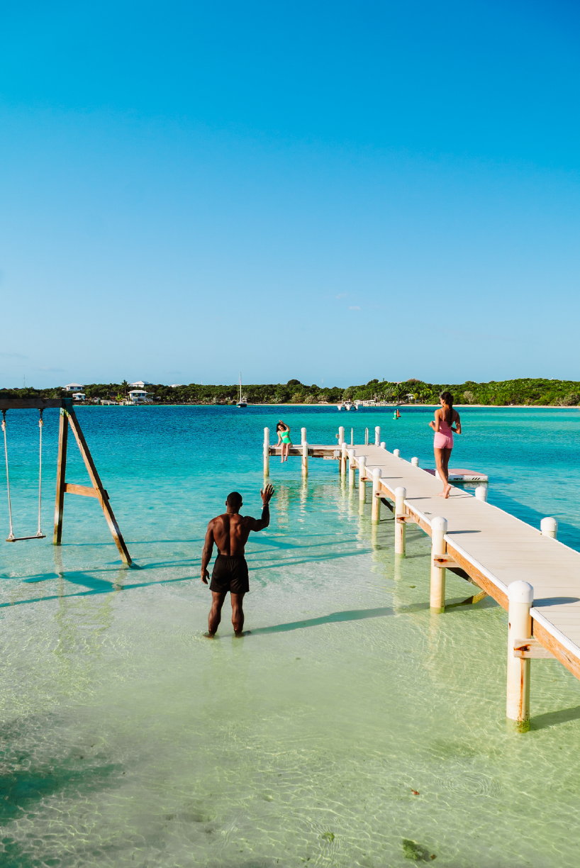 A scenic beach scene with a pier extending into clear blue water, children playing, and an adult standing in shallow water under a bright blue sky.