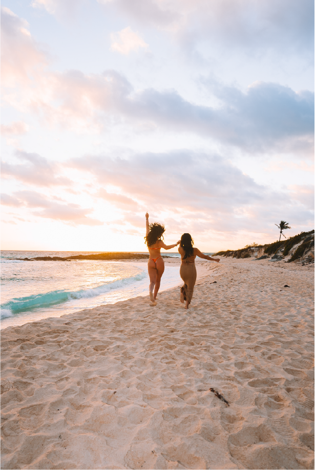 Two women walking hand in hand on a sandy beach during sunset, with one raising an arm and palm in the air, and a palm tree on the distant shoreline.
