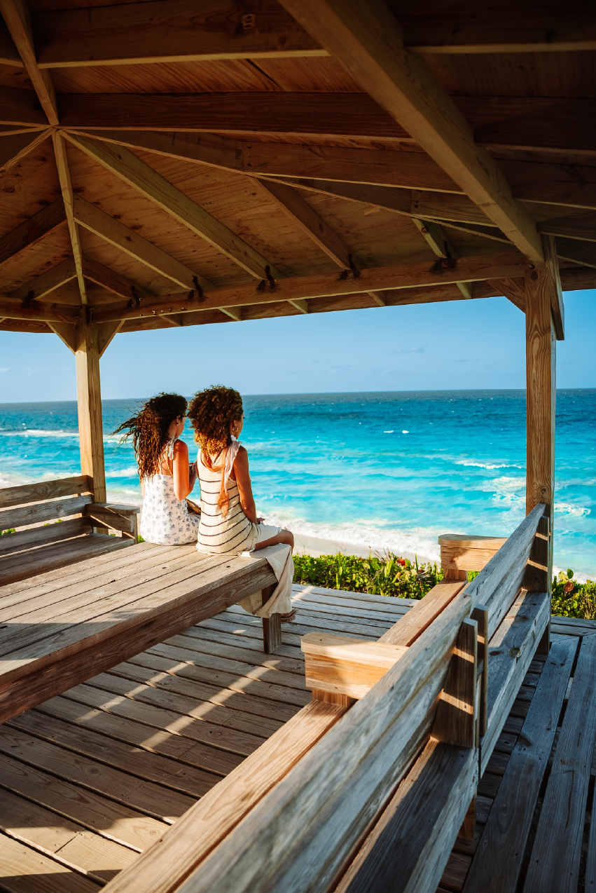 Two women sitting on a wooden bench at a beachfront gazebo, looking out at the ocean with a blue sky.