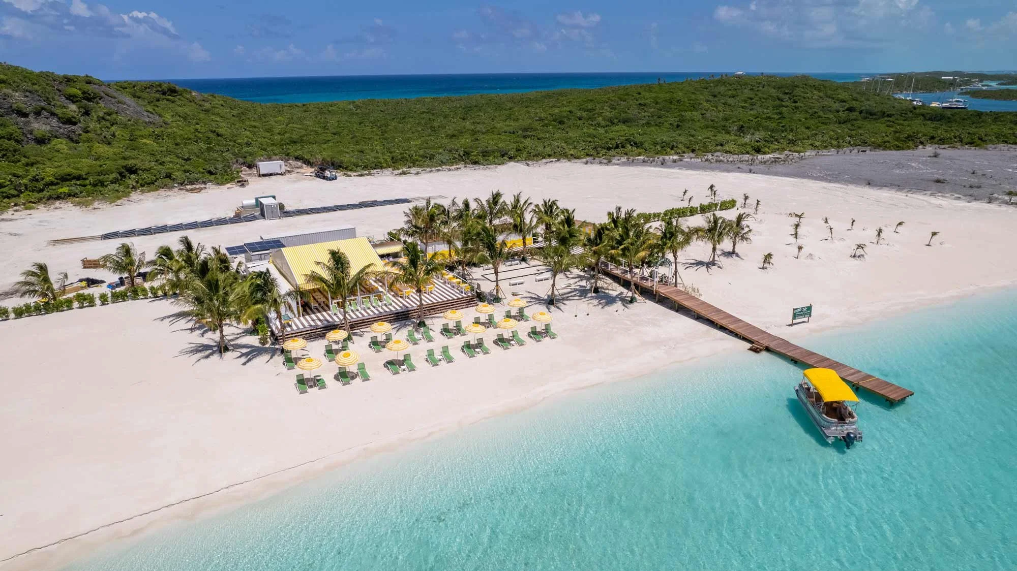 Aerial view of a beachside resort with a dock, yellow umbrellas, and lounge chairs, surrounded by turquoise water, palm trees, and lush green hills in the background.