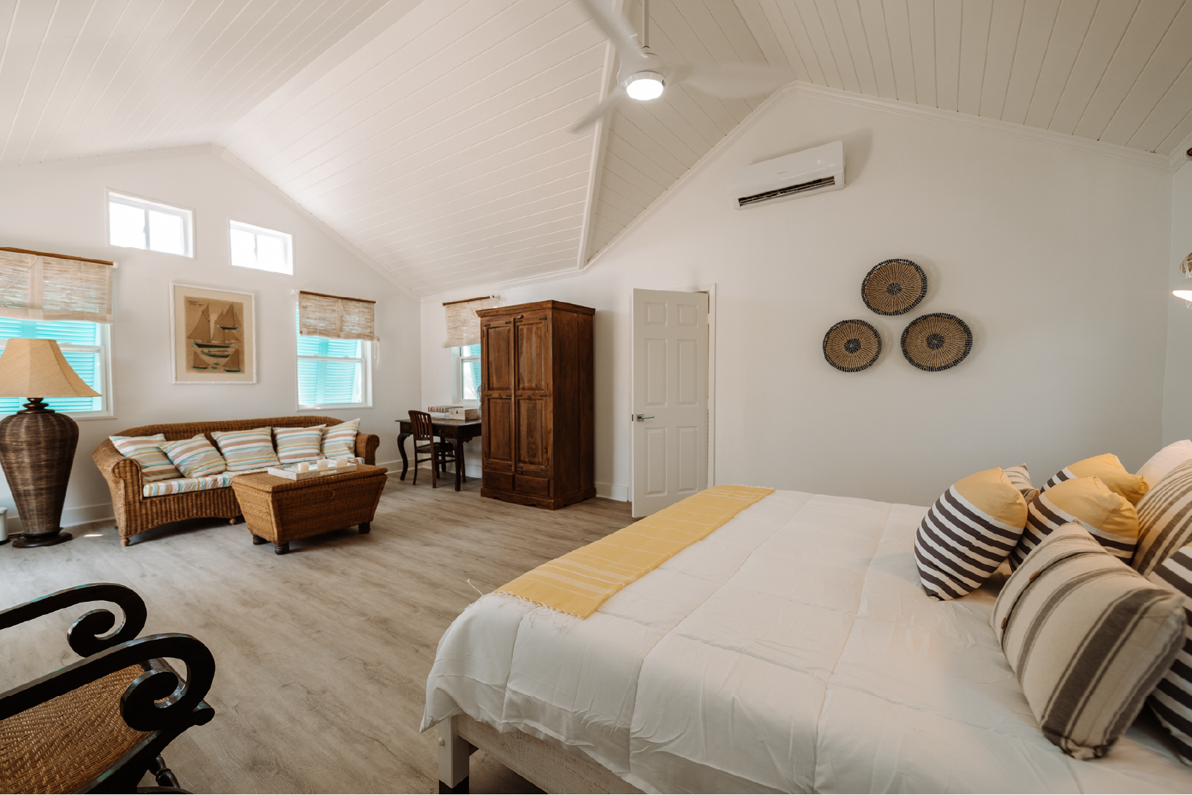A bedroom with a high, vaulted ceiling painted white, featuring a ceiling fan and an air conditioning unit. The room has four small windows near the ceiling, with three at the top and two with window coverings. The room includes a bed with striped pi