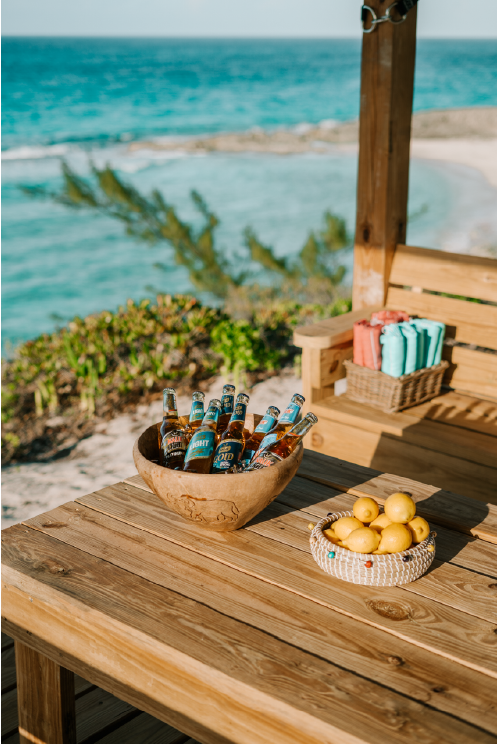 Wooden outdoor table on a beach deck with a bowl of bottled beer, a small bowl of lemons, and pastel-colored napkins in a basket, overlooking the ocean.