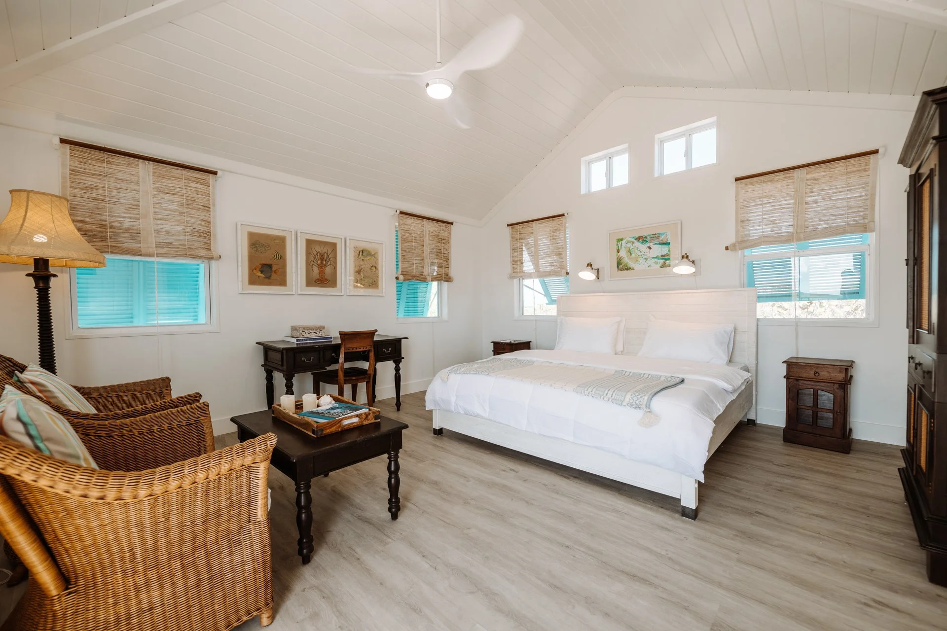Bright, airy beach-themed bedroom with white walls, wooden floors, a large white bed, wicker chairs, and multiple windows with woven shades.