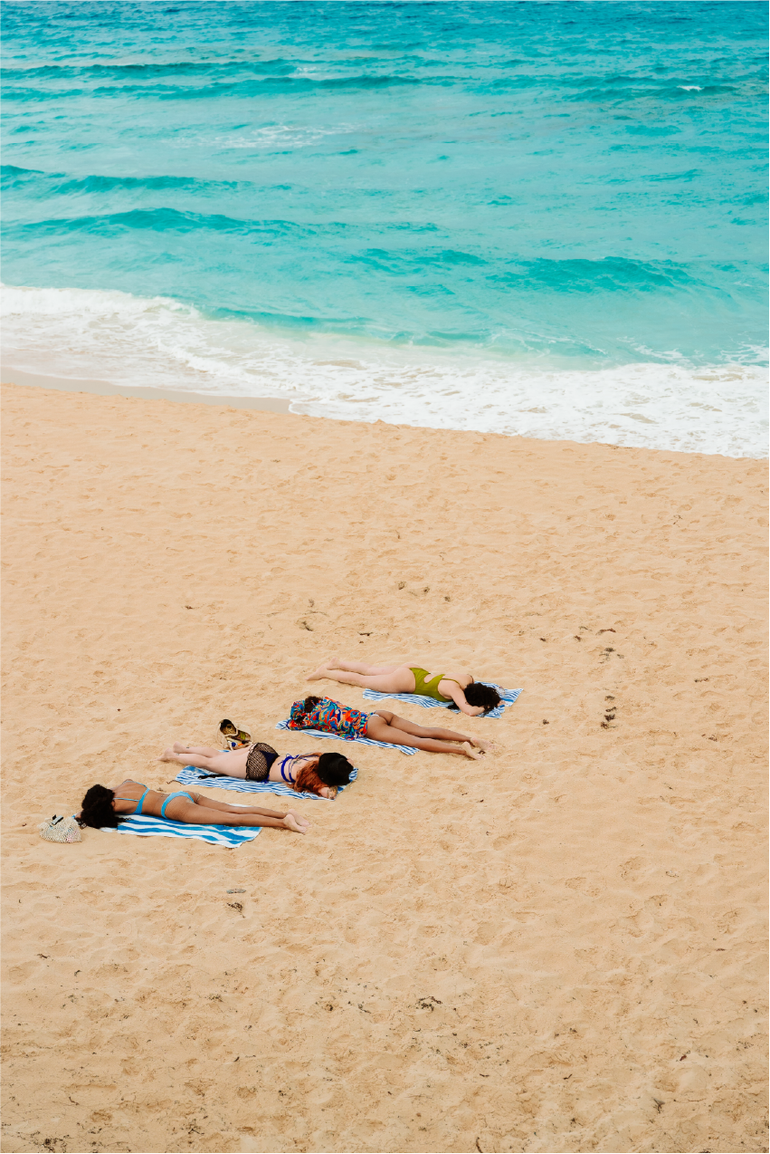 Three women lying on beach towels on sandy beach near ocean waves, sunbathing under clear skies.
