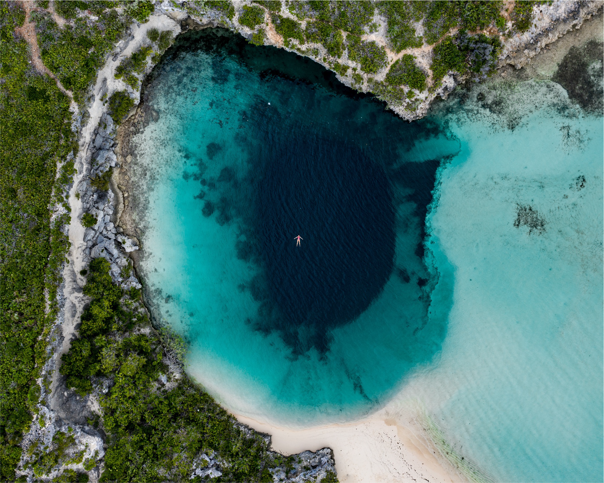 Aerial view of a person floating on a boat in a turquoise coastal bay surrounded by rocky cliffs and green vegetation.