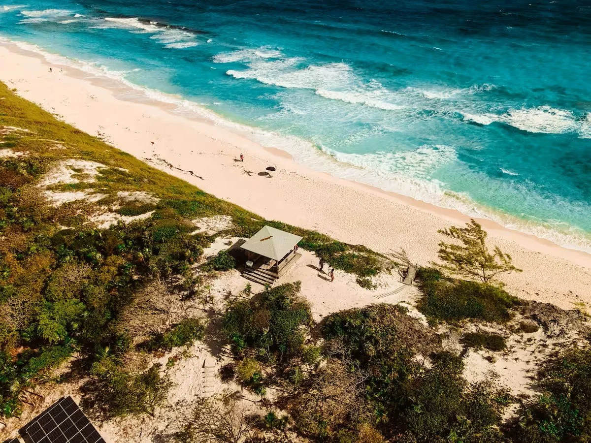 Aerial view of a beach with sand dunes, green vegetation, a small wooden shelter, and ocean waves