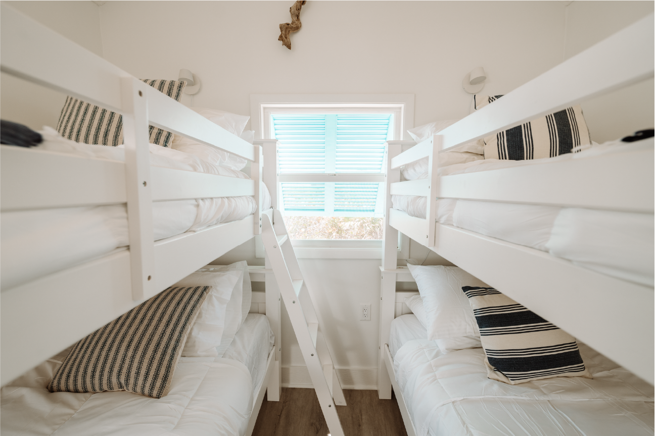 Empty bedroom with two white bunk beds, striped pillows, a window with blue blinds, hardwood flooring, and wall-mounted reading lights.