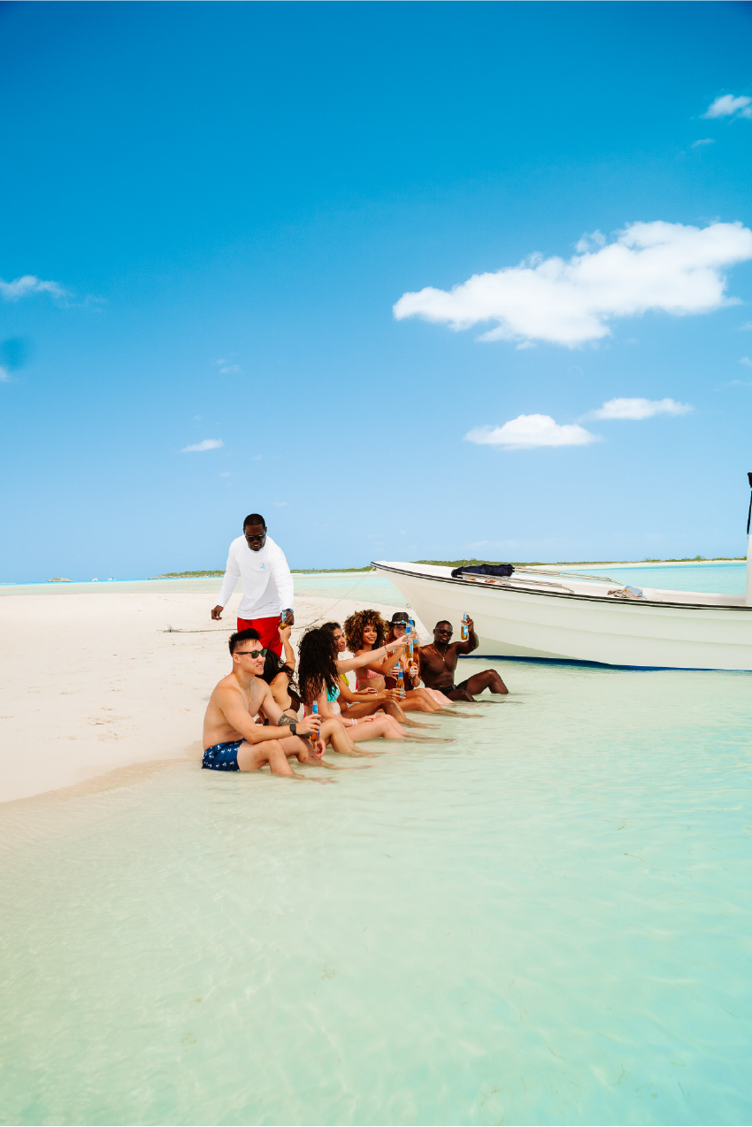 Group of friends sitting in shallow water on a beach, raising bottles in a toast, with a boat nearby under a blue sky with some clouds.