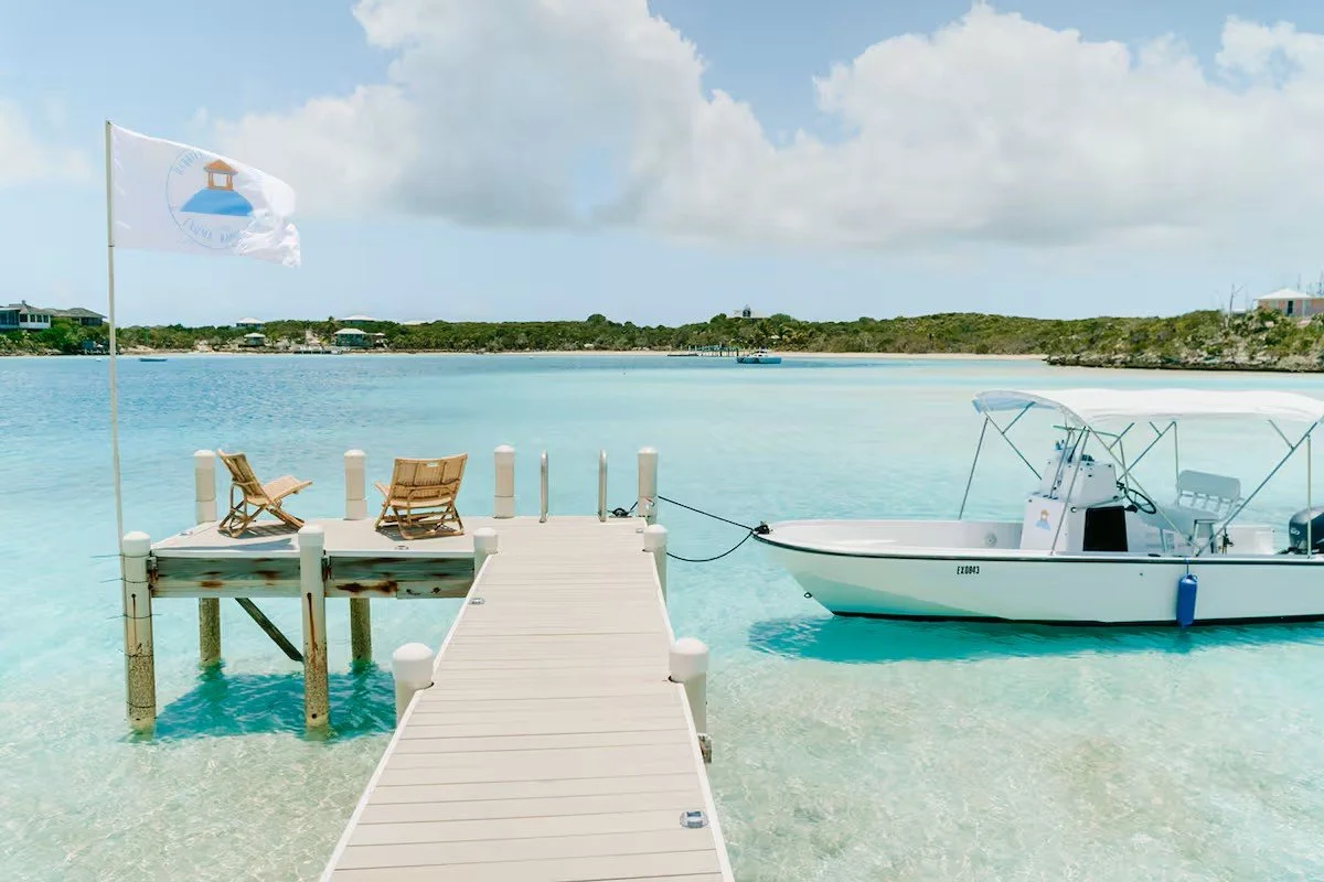 A wooden dock extending over clear turquoise water with two folding chairs on it, a flag with a boat logo in the wind, and a small white motorboat tied to the dock, with a distant shoreline and partly cloudy sky in the background.