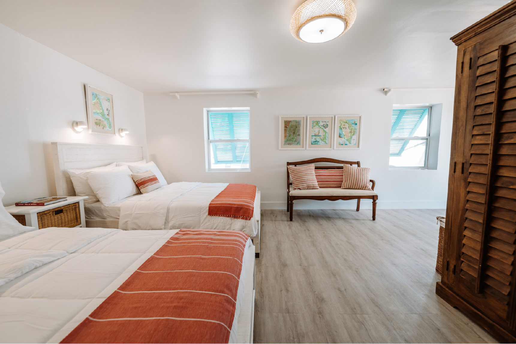 A bright bedroom with two white beds featuring red and beige throws and pillows, a wooden bench with striped pillows, a wooden wardrobe, and three framed pictures on the wall. Two windows with blue shutters let in natural light.