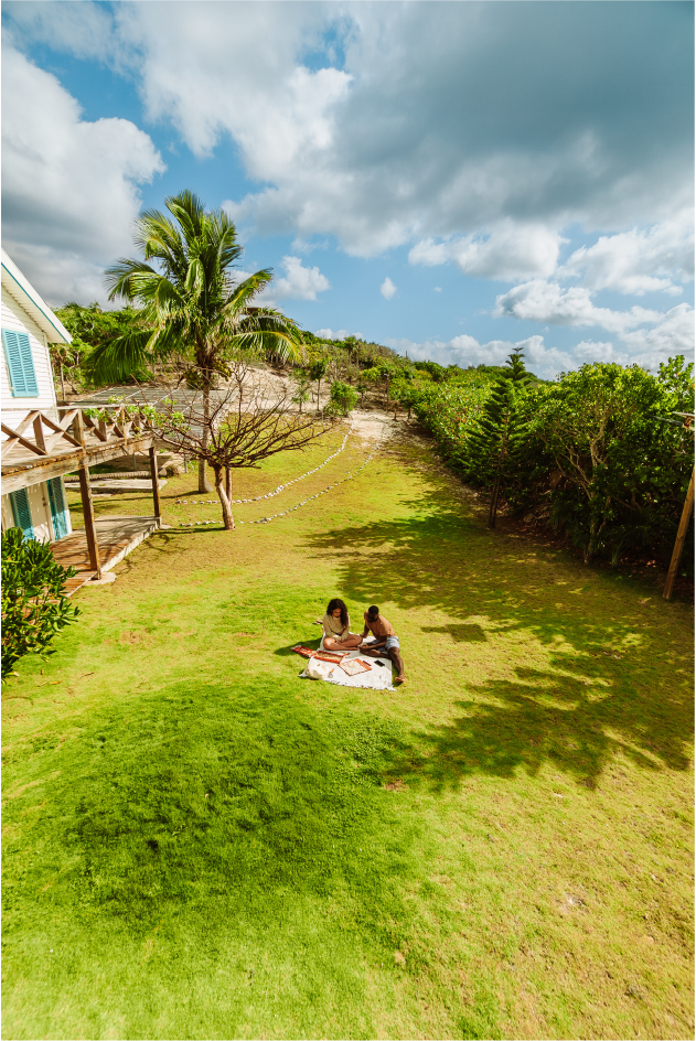 Two people sitting on a blanket on a grassy lawn, under a partly cloudy sky with tropical trees and a house in the background.