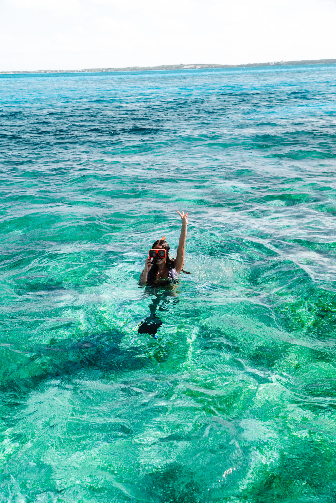 Woman in snorkeling gear waving in clear turquoise ocean water with land in the distance.