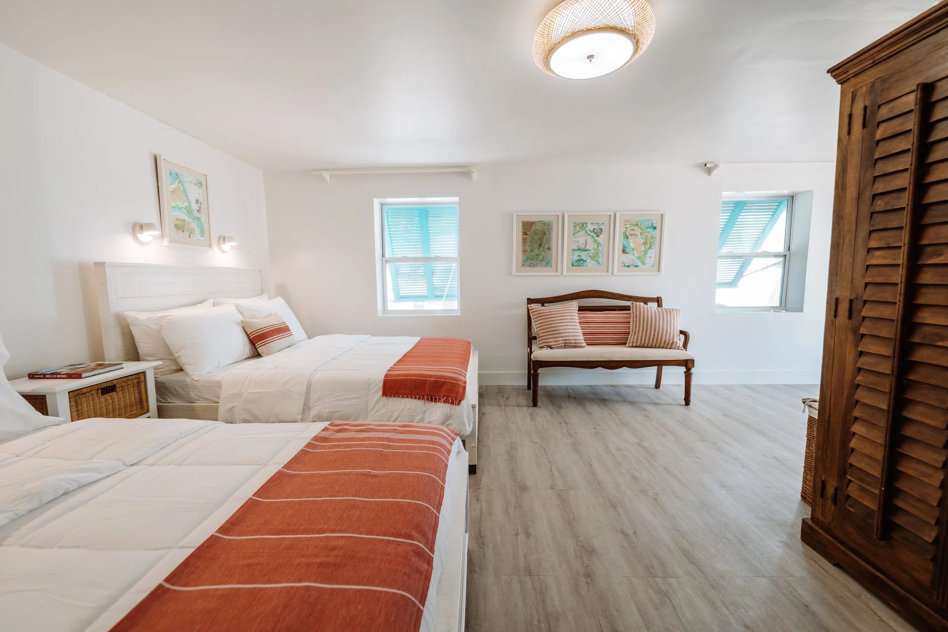 A bright bedroom with two beds covered in white bedding and orange throws, a wooden bench with striped cushions, framed artwork on the walls, and windows with teal blinds.