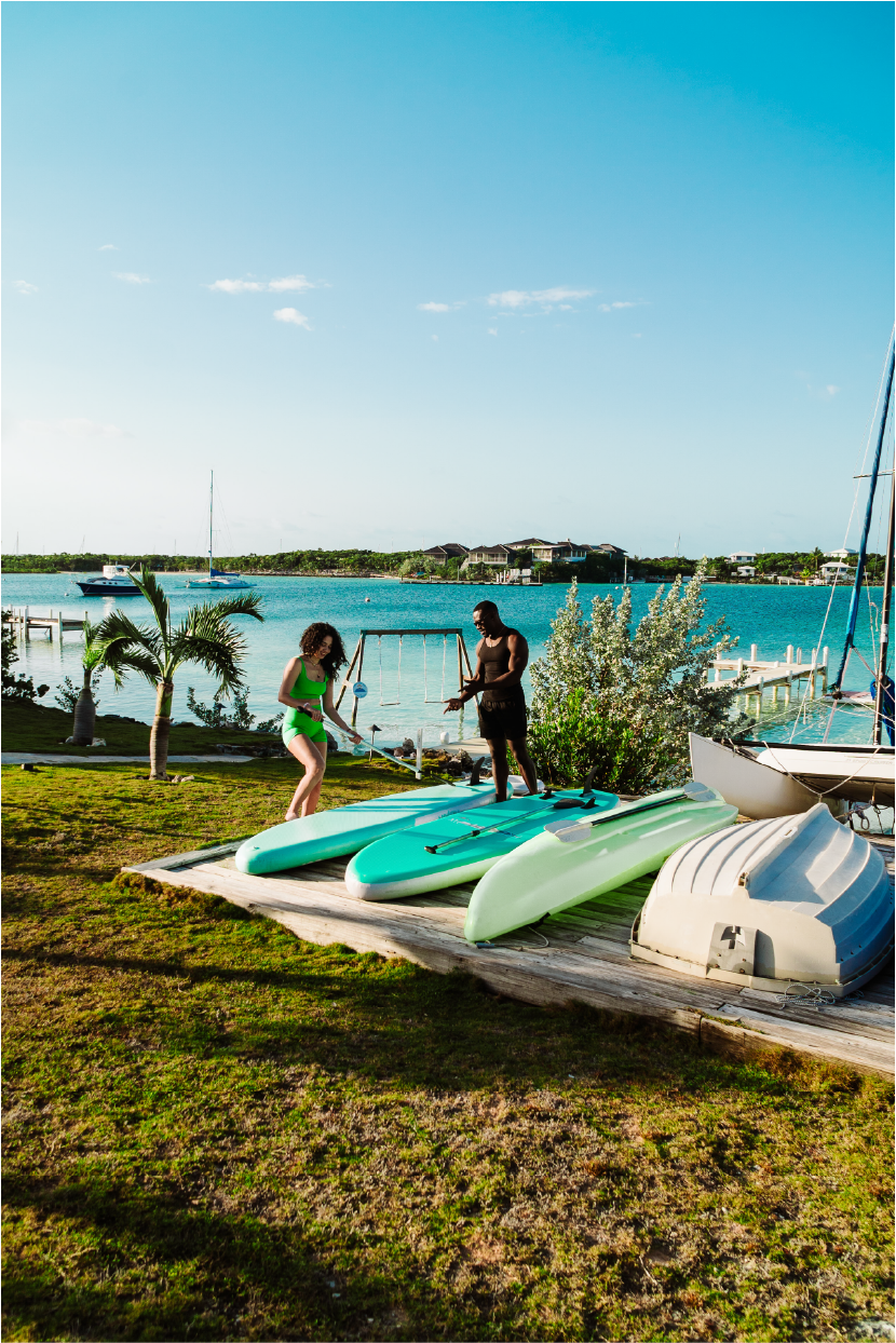 Two people preparing paddleboards near a lake under a blue sky, with sailboats anchored in the water and houses in the background.