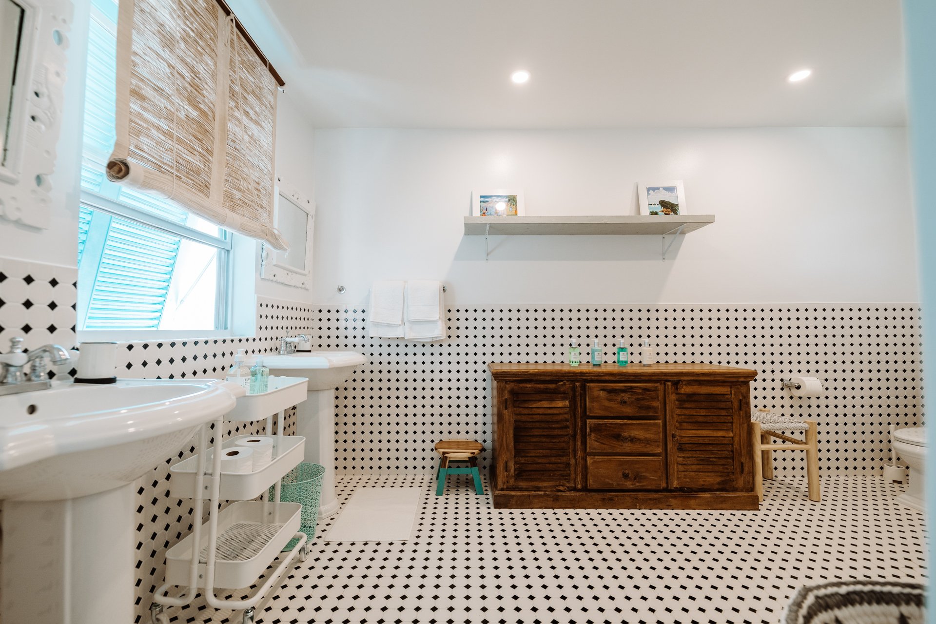 A bathroom with black and white patterned floor and wall tiles, two containing sinks, a wooden cabinet, a window with a bamboo blind, and towels on a rack.