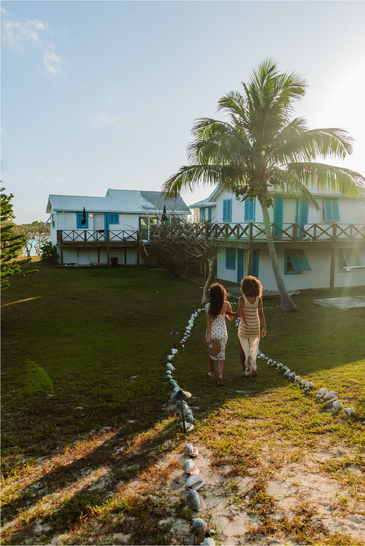 Two women walking along a stone-lined pathway towards a white beach house with blue shutters, in a tropical setting with palm trees during sunset.