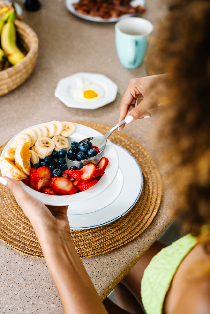 A woman holding a bowl of sliced bananas, strawberries, and blueberries, with a spoon in her hand, at a breakfast table. There is a plate with a fried egg, a mug, and a plate of bacon in the background.