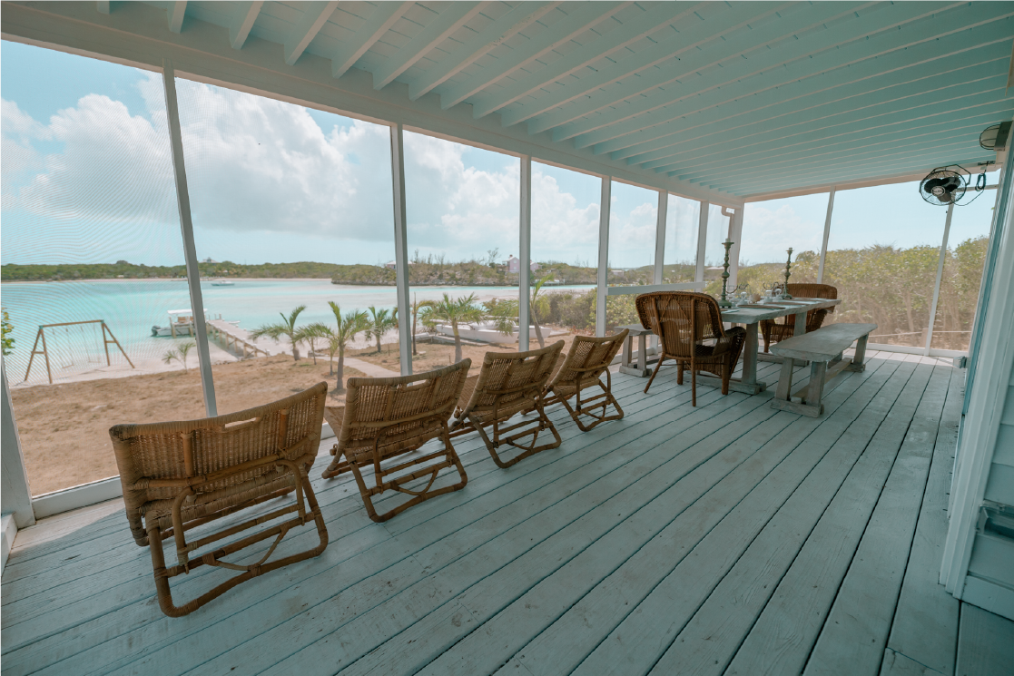 Beachside porch with a wooden table, wicker chairs, and a view of a sandy beach, palm trees, and the ocean.